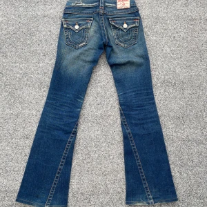 True religion jeans - Säljer dessa otroligt snygga lågmidjade klassiska blå bootcut jeans från True Religion💋De har snygga slitningar (designad så) och kontrastsömmar😻Jeansen har broderade bakfickor med knappar och orange detaljer. Midjemått 34cm, innerbenslängd 78cm, grenhöjd 17cm💕Strl 24, som XXS/XS!! De har en fläck på ena knäskålen, men inget man tänker på (har ej provat att tvätta bort, så det funkar nog) kolla sista bilden!!❤️Gammal modell=sällsynt & ovanligt. Hör av er vid funderingar eller bud🤗