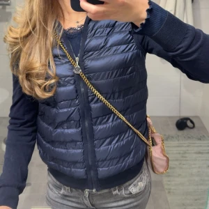 Mörkblå dunväst från Moncler - Snygg mörkblå dunväst från Moncler med quiltad framsida och ärmar i mjuk trikå. Västen har dragkedja framtill och ribbade muddar vid ärmslut och nederkant. Perfekt för lager-på-lager och en sportig look.