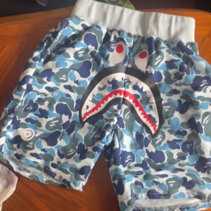Blå camo-shorts med hajmotiv från BAPE - Säljer ett par blå camo-shorts från BAPE med ikoniskt hajansikte framtill. Shortsen har bred resår i midjan och är perfekta för en streetwear-look. Färgerna går i olika blå nyanser med vita och svarta detaljer.