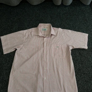 Rutig kortärmad skjorta från Lacoste - Kortärmad skjorta från Lacoste i vitt och rött rutigt mönster. Klassisk krage, bröstficka med Lacoste-logga och knäppning framtill. Skjortan är tillverkad i en blandning av polyester och bomull.
