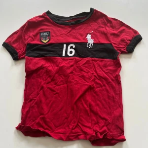 Röd t-shirt från Polo Ralph Lauren med tryck - Röd t-shirt från Polo Ralph Lauren med svart krage och svart rand över bröstet. Tryck med siffran 16, vit logga och ett  emblem på bröstet. Perfekt för dig som gillar sportig stil. För barn