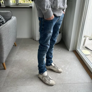 Blå jeans från Nudie Jeans - Säljer ett par klassiska blå jeans från Nudie Jeans med orangea sömmar och fem fickor. Byxorna har normal passform och raka ben. Tillverkade i denim med snygga slitningar och detaljerad bakficka.