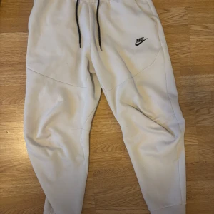 Vita mjukisbyxor från Nike - Säljer ett par vita mjukisbyxor från Nike med svart snörning i midjan och en diskret logga på benet. Byxorna har en bekväm passform och är perfekta för en avslappnad stil. De har även en praktisk ficka med dragkedja på sidan.