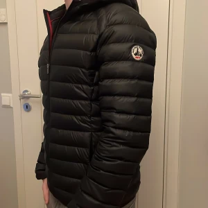 Svart pufferjacka med huva - Snygg svart pufferjacka från Jott med huva och dragkedja framtill. Jackan har ett quiltat mönster och en logotyp på ärmen. Perfekt för kyligare dagar. Nypris ligger på 2200 kr.