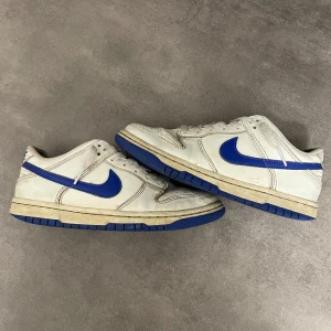 Nike Dunk i vitt och blått - Säljer ett par klassiska Nike Dunk sneakers i vitt med blå detaljer. Skorna har en rund tå och snörning framtill. Perfekta för en sportig look eller vardagsstil.