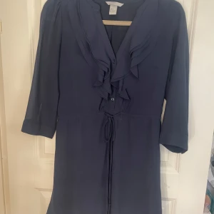 Mörkblå klänning med volangdetaljer - Elegant mörkblå klänning med volangdetaljer framtill och knytband i midjan. Klänningen har trekvartsärmar och en v-ringad design som ger en stilren look. Perfekt för en chic och sofistikerad stil.