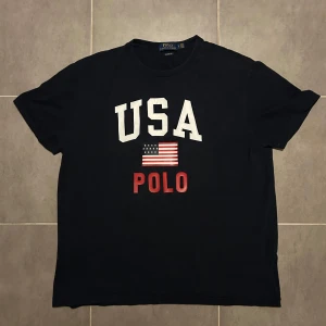 Vintage Ralph Lauren USA T-shirt Navy L - Snygg mörkblå vintage t-shirt från Ralph Lauren med tryck av 'USA' och en amerikansk flagga i vitt och rött. Texten 'POLO' i rött under flaggan. Perfekt för en casual look.