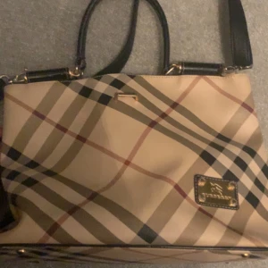 Rutig handväska från Burberry - Säljer en stilren rutig handväska från Burberry med  mönster. Väskan har svarta handtag och detaljer i guld. Insidan är fodrad med Burberry-logotyp och har en praktisk dragkedja.