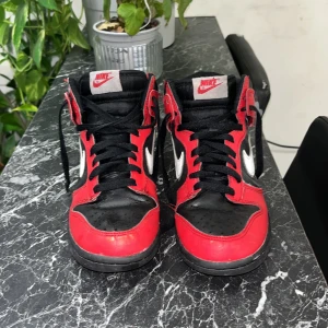 Nike sneakers i rött och svart - Snygga Nike sneakers i en klassisk röd och svart färgkombination med vita detaljer. Skorna har hög snörning och en ikonisk Nike-logga på sidan. Perfekta för dig som vill ha en stilren och sportig look.