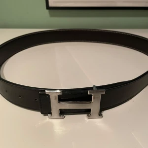 Svart bälte med silverfärgat spänne - Snyggt svart bälte med ett stort, silverfärgat H-format spänne. Perfekt för att ge en stilren touch till din outfit. Bältet har en klassisk design och passar till många olika stilar.
