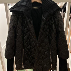 Svart quiltad jacka med dragkedja - Snygg svart quiltad jacka med dragkedja och knappar. Jackan har en hög ribbad krage och långa ärmar med ribbade muddar. Perfekt för kyligare dagar.