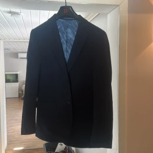 mörkblå kavaj - Elegant mörkblå kavaj med klassisk design. Kavajen har två knappar framtill och är helfodrad med ett ljusblått innerfoder. Perfekt för formella tillfällen. Storleken är 48 regular slim fit 