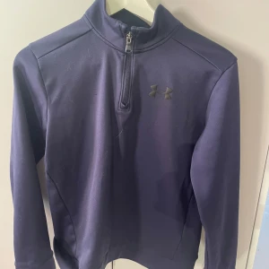 Mörkblå tröja från Under Armour half zip  - Säljer en mörkblå tröja från Under Armour med halv dragkedja och logotyp på bröstet. Tröjan har en lös passform och är perfekt för träning eller vardagsbruk. Tillverkad i ett mjukt och bekvämt material. Ca stl 152