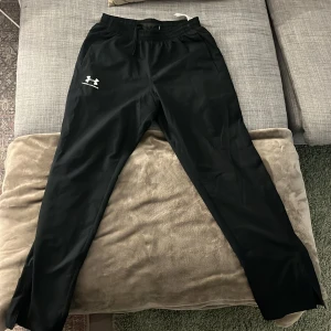 Svarta träningsbyxor från Under Armour - Säljer ett par svarta träningsbyxor från Under Armour. Byxorna har en elastisk midja och är i en loose passform, perfekt för träning. De är tillverkade i ett lätt syntetmaterial och har Under Armour-loggan tryckt på benet. Längst ner vid foten finns det en dragkedja på båda ben för att anpassa passformen runt över foten. Det är 10/10 skick och ny pris 599kr mitt pris 400 kan sänkas som lägst 350