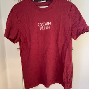 Vinröd T-shirt från Calvin Klein! - Vinröd T-shirt från Calvin Klein i storlek M. T-shirten har ett tryck på bröstet samt på armen.  Obs Bokstaven C på armtrycket är löst, därav priset. Fint skick.