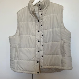 Beige dunväst med knappar - Säljer en  beige dunväst med knappar framtill. Västen har en hög krage och två sidofickor. Passar bra till lager-på-lager-stil. Stl är M/L
