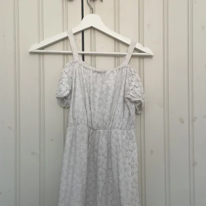 Vit spetsklänning med axelband - Charmig vit långklänning i spets med axelband och korta ärmar. Perfekt för sommarens festligheter. Klänningen har en elastisk midja och ett vackert blommönster i spetsen. En fläck som finns som troligtvis går att tvätta bort.