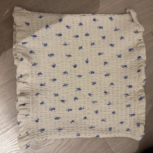 Vit tubtopp med blå blommor - Charmig vit topp med små blå blommor och volangdetaljer. Toppen har en smockad design som ger en elastisk och bekväm passform. Perfekt för en somrig look! 