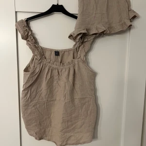 Set med volangdetaljer - Säljer en söt beige linnetopp med tillhörande shorts från shein med volangdetaljer vid axlarna. Toppen är ärmlös och har en luftig passform, perfekt för varma dagar. Materialet är mjukt och bekvämt.