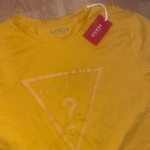 Gul t-shirt från Guess - Säljer en snygg gul t-shirt från Guess med det ikoniska triangellogotypen tryckt på framsidan. T-shirten har en klassisk rund hals och korta ärmar. Perfekt för en avslappnad stil.