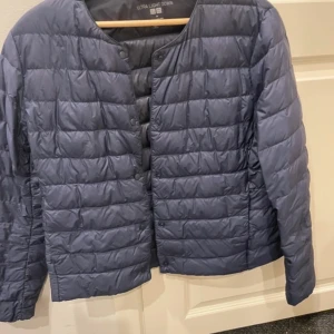 Uniqlo dunjacka - Säljer en stilren blå ultralätt dunjacka från Uniqlo. Jackan har en quiltad design med knappar framtill och långa ärmar. Perfekt för lager-på-lager under kyliga dagar. 