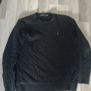 Svart stickad tröja från Ralph Lauren - Säljer en stilren svart stickad tröja från Ralph Lauren med kabelstickat mönster. Tröjan har långa ärmar och en rund halsringning. Den ikoniska röda logotypen är broderad på bröstet. Perfekt för en klassisk look.