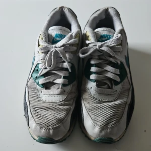 Nike Air Max sneakers i vitt och turkos - Säljer ett par klassiska Nike Air Max sneakers i vitt med turkosa och svarta detaljer. Skorna har den ikoniska Air Max-sulan och snörning framtill. Perfekta för en sportig look.