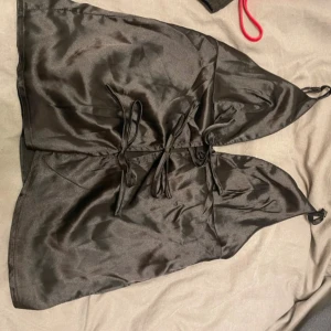 Svart satinlinne med knytband - Elegant svart satinlinne med knytband framtill. Toppen har en djup v-ringning och tunna axelband, vilket ger en stilren look. Perfekt för en kväll ute eller en speciell tillställning.