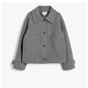 Grå kappa - Snygg grå ulljacka med en klassisk krage och stora framfickor. Jackan har knappar framtill och långa ärmar med knappar vid ärmsluten. Perfekt för kyligare dagar.