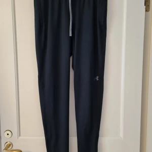 Svarta träningsbyxor från Under Armour - Snygga svarta träningsbyxor från Under Armour med elastisk midja och dragsko. Byxorna har en smal passform och är perfekta för träning. De har även praktiska fickor på sidorna. Storlek M. Kan hämtas i Vallentuna.