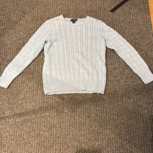 Ljusblå stickad tröja från Polo Ralph Lauren - Säljer en stilren ljusblå stickad tröja från Polo Ralph Lauren med kabelstickat mönster i storlek 14-16 som motsvarar XS. Tröjan har långa ärmar och en rund halsringning. Den ikoniska loggan är broderad i gult på bröstet. Perfekt för en klassisk och avslappnad look. Nypris 1249 mitt pris 399 först till kvarn 