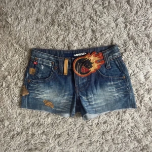 Jeansshorts från Miss Sixty - Snygga jeansshorts från Miss Sixty med coola detaljer som en stor broderad patch i eldflames-design. Shortsen har en klassisk femficksmodell och är lågmidjade.  Storlek 24, 35 cm i midjemått❤️
