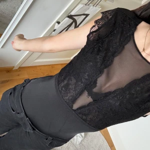 Svart spetsbody - Elegant svart body med spetsdetaljer och transparenta partier. Blusen har korta ärmar och en djup v-ringning, vilket ger en sofistikerad look. Perfekt för en kväll ute eller en speciell tillställning.