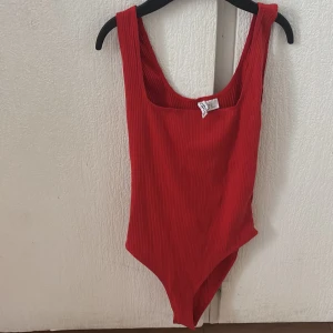 Röd ribbad bodysuit från H&M - Snygg röd ribbad bodysuit från H&M med djup urringning. Perfekt för att styla med jeans eller kjol för en trendig look. Passar utmärkt för både vardag och fest.