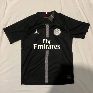 Svart PSG fotbollströja med Fly Emirates - Snygg svart PSG fotbollströja med Fly Emirates-logga och Jordan-märke. Tröjan har korta ärmar och ett stilrent mönster i vitt och grått. Perfekt för fotbollsfans som vill ha en cool look på planen eller på stan.