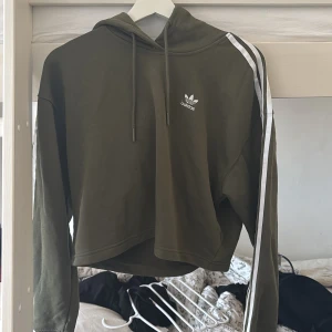 Grön hoodie från Adidas - Säljer en snygg grön hoodie från Adidas med vita ränder på ärmarna och logga på bröstet. Hoodien har en croppad passform och dragsko i huvan. Perfekt för en avslappnad stil.