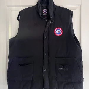 Svart dunväst från Canada Goose - Säljer en svart dunväst från Canada Goose med ett broderat märke på bröstet. Västen har knappar och två stora fickor framtill. Perfekt för kyliga dagar. Har liten skada på axeln men ser knappt.