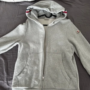 Moncler Hoodie - Säljer en stilren grå huvtröja från Moncler med dragkedja och broderad logga på ärmen. Tröjan har en bekväm passform och är perfekt för kyligare dagar. Huvan har en snygg randig detalj i rött och vitt.