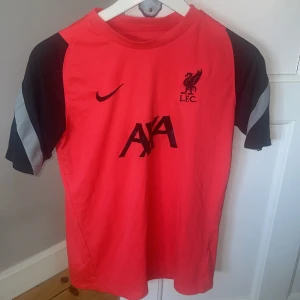 Röd fotbollströja från Nike - Säljer en röd fotbollströja från Nike med svarta och grå detaljer. Tröjan har korta ärmar och L.F.C.-logga på bröstet. Perfekt för fotbollsfans!