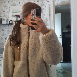 Beige teddyjacka med dragkedja - Mysig beige teddyjacka med dragkedja framtill. Perfekt för kyliga dagar med sin fluffiga och varma design. Jackan har långa ärmar och en hög krage för extra värme.🌸