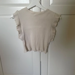 Beige ribbad topp från Zara - Säljer en söt beige ribbad topp från Zara. Toppen är ärmlös med volangdetaljer vid axlarna och har en croppad passform. Perfekt för varma dagar!