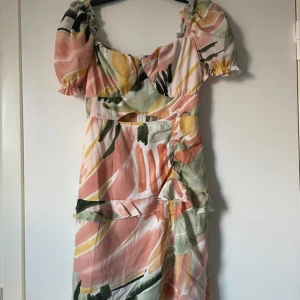 Färgglad klänning med volanger - Säljer en färgglad klänning med abstrakt mönster i rosa, grön, gul och svart. Klänningen har korta puffärmar och volangdetaljer framtill. Perfekt för sommarens fester!