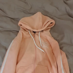 Rosa adidas hoodie med dragkedja - Säljer en mysig rosa hoodie med dragkedja och vita snören. Den har en luva och vita ränder längs ärmarna. Perfekt för en avslappnad stil.