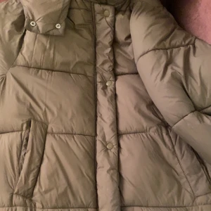 Olivgrön extra lång Jacka - Säljer en olivgrön pufferjacka från Ginatricot. Jackan har en pufferdesign med långa ärmar och knappar framtill. Perfekt för kyliga dagar med sin varma och bekväma passform.