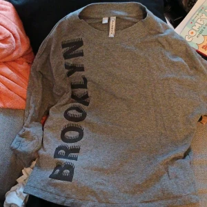 Grå tröja från Rainbow med text - Säljer en grå tröja från Rainbow med texten 'BROOKLYN' i svart på framsidan. Tröjan har långa ärmar och en rund halsringning. Perfekt för en avslappnad stil.