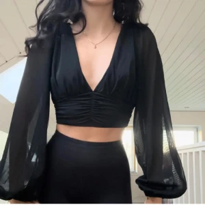 Svart v-ringad croppad blus - Snygg svart blus med v-ringning och långa, transparenta ärmar. Den är croppad och har en åtsittande passform med rynkdetaljer framtill. Perfekt för en stilren look.