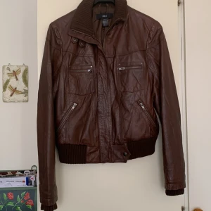 Brun skinnjacka från Ellos - Säljer en snygg brun skinnjacka från Ellos med dragkedja och flera praktiska fickor. Jackan har en ribbad krage och muddar, vilket ger en cool look. Perfekt för höst och vår!