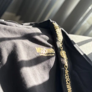 Svart tröja från Moschino - Snygg svart tröja från Moschino med guldfärgad logotyp och detaljer. Tröjan har en rund halsringning och är dekorerad med ett band med Moschino-logotypen längs ärmarna. Perfekt för en stilren look. Kvitto finns.