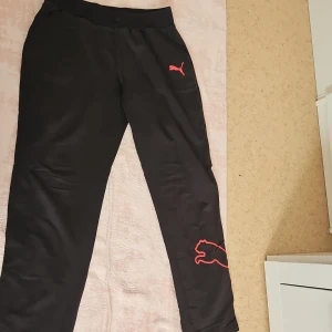 Svarta mjukisbyxor från Puma - Säljer ett par svarta mjukisbyxor från Puma med röd logga på benet och vid fickan. Byxorna har en elastisk midja för extra komfort. Perfekta för en avslappnad stil.