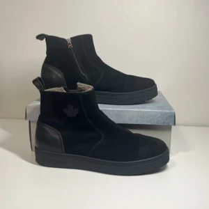 Svarta boots från Canada Snow - Snygga svarta boots från Canada Snow med dragkedja på sidan och en stilren design. Skorna har en bekväm ullfodring som håller fötterna varma. Perfekta för kyligare dagar med en diskret lövdetalj på sidan.